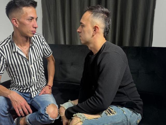 Two BigDickTop69s, sitting on a black couch, facing each other. One wears a striped shirt and ripped jeans, the other a black sweater and torn jeans.