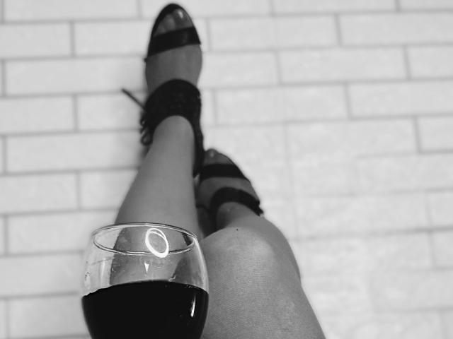 Black and white photo. VittoriaI's legs extend diagonally from bottom right to top left. She wears sheer stockings with dark straps. Her right foot is in a black open-toe sandal with a strap. A glass of dark red wine is in her left hand, positioned near her knee. The background is a white brick wall. The composition emphasizes contrast between her legs and the glass. Simple, focused, and minimalistic.