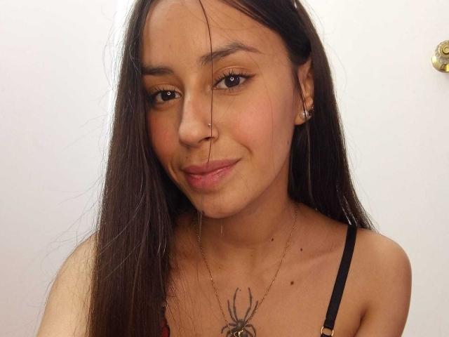 LizzyGrey with long brown hair, nose ring, earrings, and a spider necklace on black strap, smiling softly, has a small fly on her face.