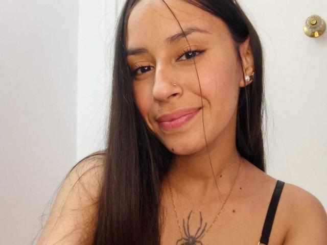 LizzyGrey has long black hair, a small nose ring, and a smile with pink lips. She's wearing a black top, showing a spider tattoo on her chest.