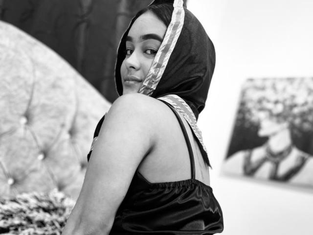 Black-and-white photo of NohemiZahar with dark hair and a headscarf, looking over her shoulder, wearing a shiny black dress, showing her bare back and arm.