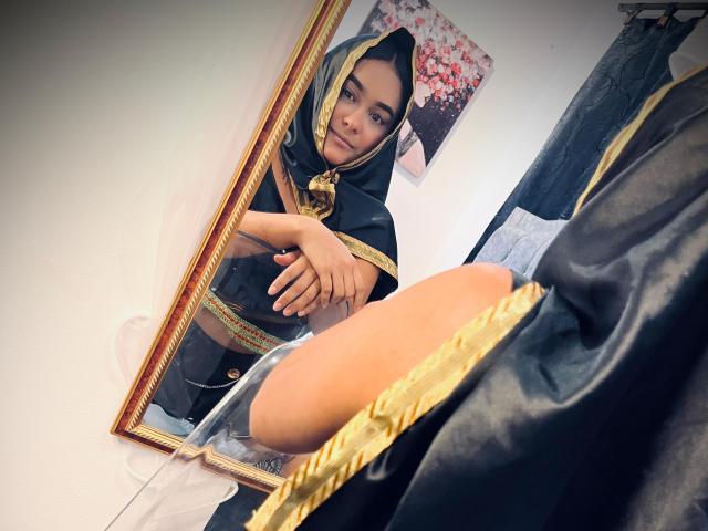 NohemiZahar with a black and gold headscarf, looking in a mirror, arms crossed, showing her bare, curvy ass reflected in the mirror.