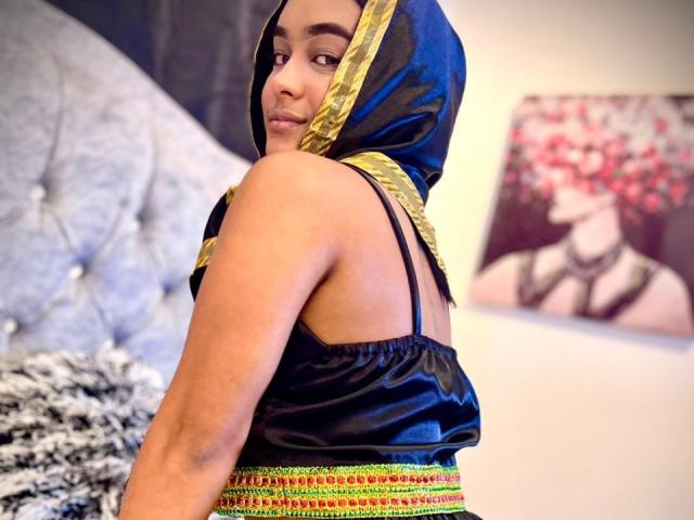NohemiZahar with brown skin, wearing a black and gold headscarf, black strap top, and colorful skirt, showing her back and looking over her shoulder. Her ass is visible.
