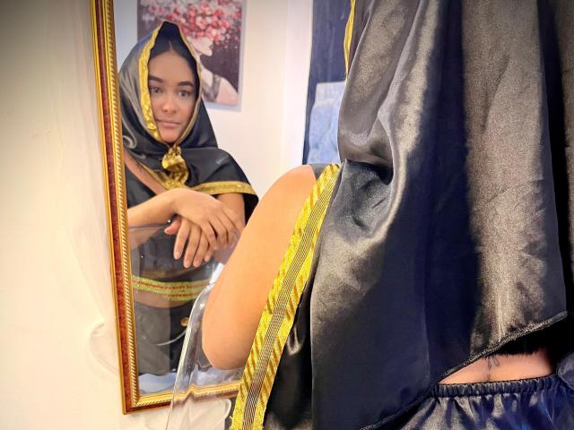 NohemiZahar's reflection shows her in a black and gold satin hooded robe, with one leg raised, exposing her bare thigh and a small buttcheek. Mirror on left.