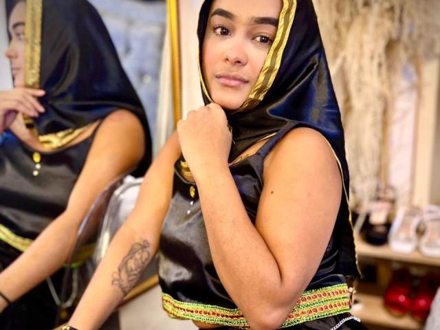 NohemiZahar with medium tits, wearing a black and gold headscarf, sleeveless top, holding her chin, reflecting in a mirror, showing a tattoo on her right arm.