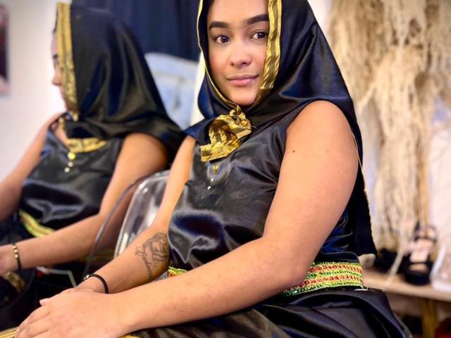 NohemiZahar with medium brown skin, dark eyebrows, and a tattoo on her left forearm, wearing a black and gold headscarf, black dress with green trim, and sitting.