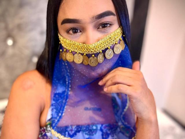 NohemiZahar with blue see-through veil, gold coin necklace, and dark hair, covering her sexy face, showing her brown eyes and slightly raised ass-cheek. Her hand touches her chin.