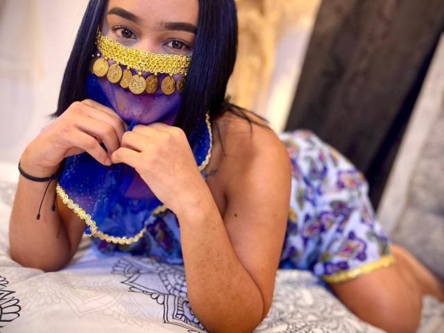 NohemiZahar with blue face mask, gold coins, and long black hair, wearing floral dress, on bed, hands near mouth, looking at camera, showing small tattoo on left arm.