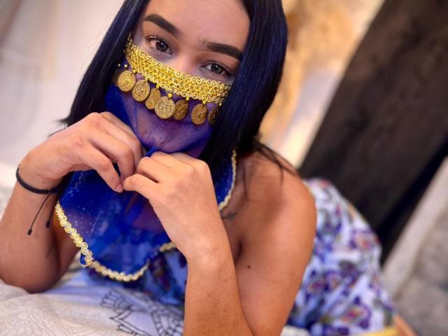 NohemiZahar with dark blue hair, wearing a gold and purple face mask, holding the mask with her hands, showing her brown eyes, and a tattoo on her right shoulder.