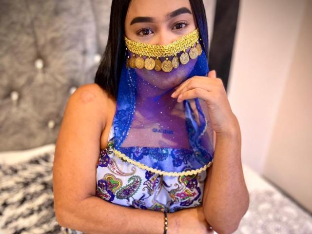 NohemiZahar with brown skin, wearing a blue sheer mask with gold coins, and a colorful patterned top, covering her tits, with black hair and a bracelet on her right wrist.