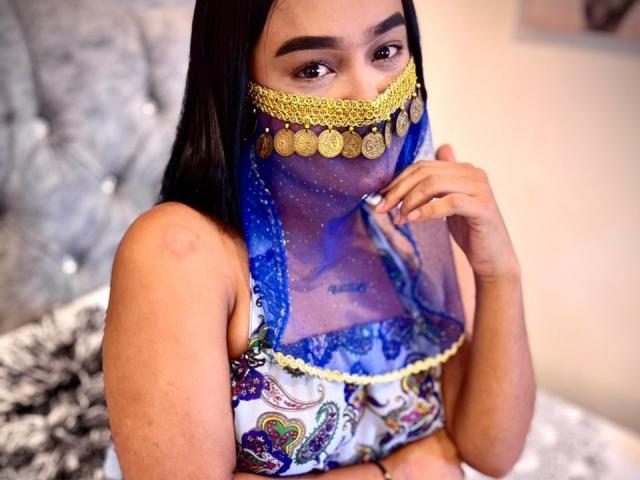 NohemiZahar with dark hair, brown skin, wearing a blue and gold patterned mask covering her nose and mouth, and a colorful, patterned top, with a hand near her mouth.