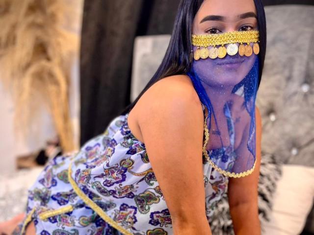 NohemiZahar with long black hair, wearing a blue and gold mask, colorful dress, and patterned fabric, kneeling on a bed, showing her ass and looking at the camera.