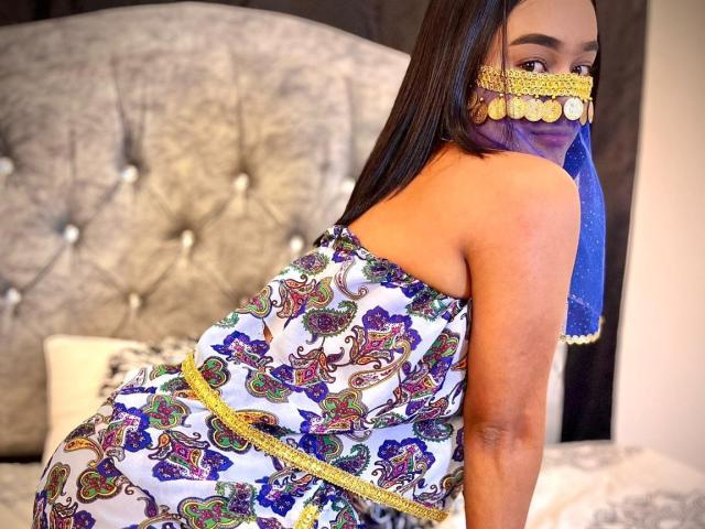 NohemiZahar has brown skin, long black hair, wearing a colorful, patterned dress, and a yellow, purple, and blue face mask, kneeling on a bed with a grey headboard.