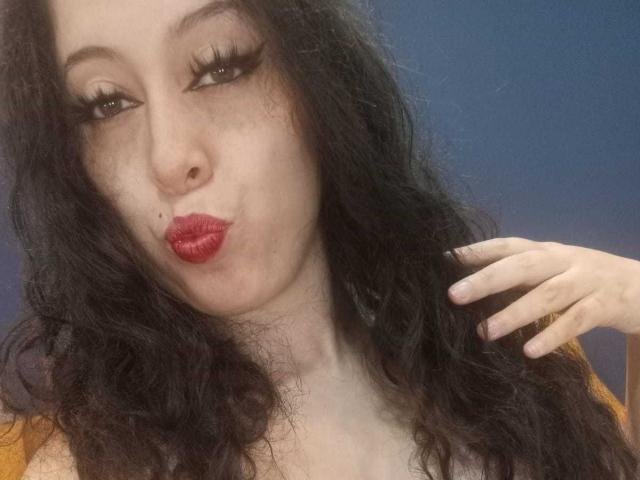KendraSimons with pouty red lips, long black curly hair, and thick eyelashes, touching her hair with a fair skin tone, brown eyes, and a small beauty mark on her cheek.