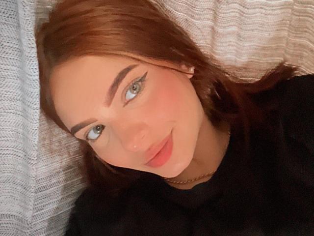 MaddieGrace with red hair, green eyes, and eyeliner, lying on a pillow, wearing a black top, smiling softly, looking to the side, with a slight blush on her cheeks.