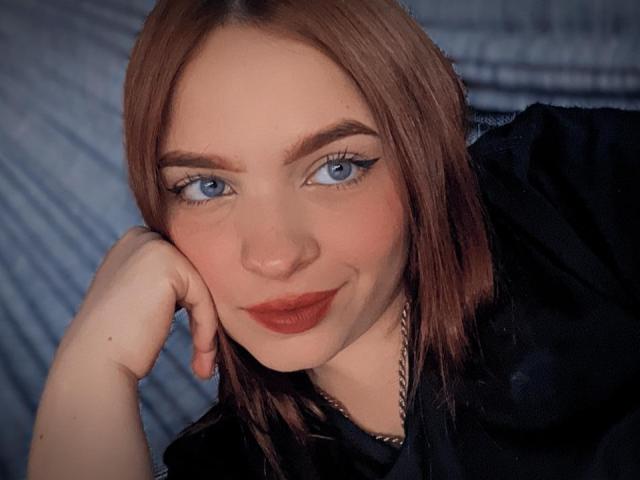 MaddieGrace has blue eyes, red lipstick, and brown hair, resting her head on her hand, wearing a black shirt, with a subtle smile, looking directly at the camera.