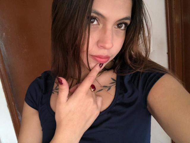 NyraVanel, com cabelos castanhos longos, olhos escuros e batom vermelho, tocando o dedo nos lábios, vestindo uma camisa azul marinho, mostrando uma tatuagem no peito..