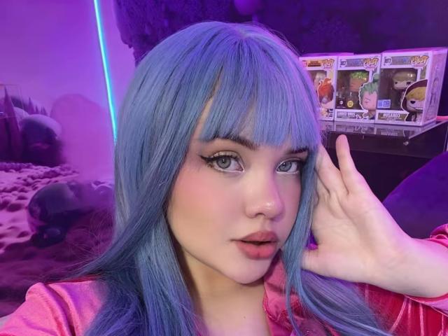 AlayaWillow with blue hair, wearing a pink shirt, has her hand near her face, showing off her eyeliner and pink lips. She's looking at the camera with a slight pout.