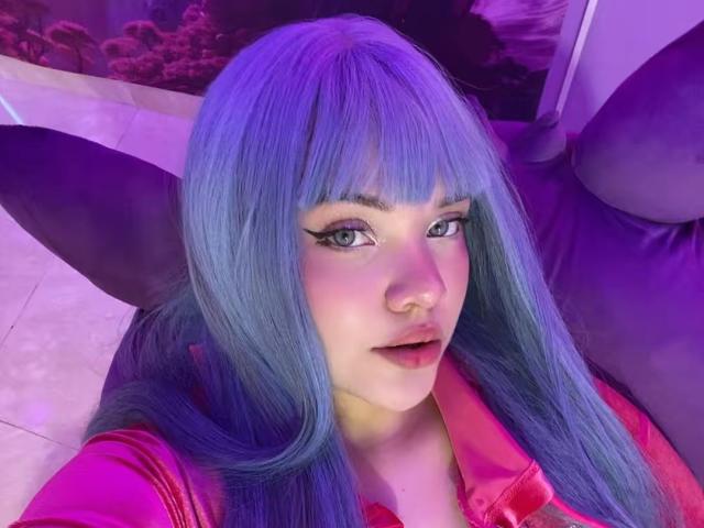 AlayaWillow with blue hair, bangs, and pink eyeliner, wearing a pink shirt, has a nose ring and purple eyes, sitting on a purple couch, looking at the camera.