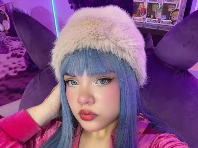 AlayaWillow with blue hair, wearing a fluffy white hat, pink jacket, and red lipstick, has her hand in her hair, staring at the camera with a pouty expression.