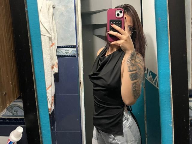 NyraVanel with brown hair, black sleeveless top, and gray shorts, holding a pink phone, visible tattoo on right arm, taking a mirror selfie in a bathroom.