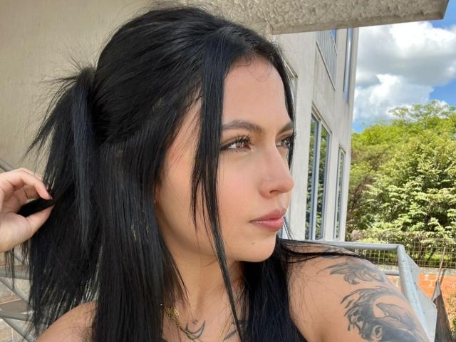 NyraVanel with black pigtails, brown eyes, and a tattoo on her right arm, looking to the side. She has light skin and a small nose. Her lips are slightly parted.