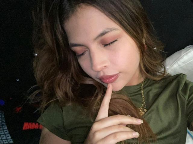NyraVanel, con cabello castaño, ojos cerrados y sombra de ojos rosa, tocando suavemente sus labios con un dedo, vistiendo una camisa verde y un collar dorado, luciendo linda y relajada..