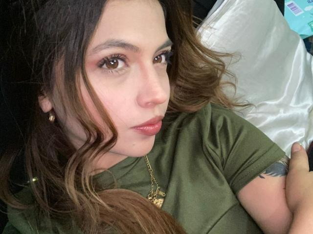 NyraVanel has wavy brown hair, green eyes, and a slightly parted pink lip. She's wearing a green shirt, gold necklace, and small gold earrings, with a tattoo on her arm.