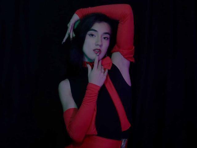 SofiaMonseur with dark hair, red and black outfit, touching her tongue to a red ring, arm raised, looking seductive, dark background, soft lighting, feminine, sexy.