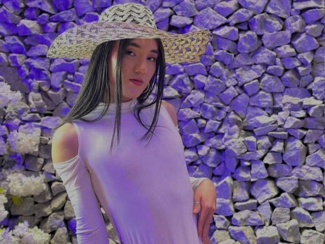 SofiaMonseur with long black hair, wearing a white, sleeveless, cut-out dress and straw hat, standing with a slight smile, purple lighting, rocks in background. Boobs visible through dress.