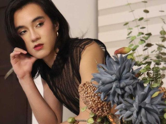 SofiaMonseur with dark hair, red lipstick, and black sheer top, leaning on blue succulents and bead-covered arm, looking slightly bored.