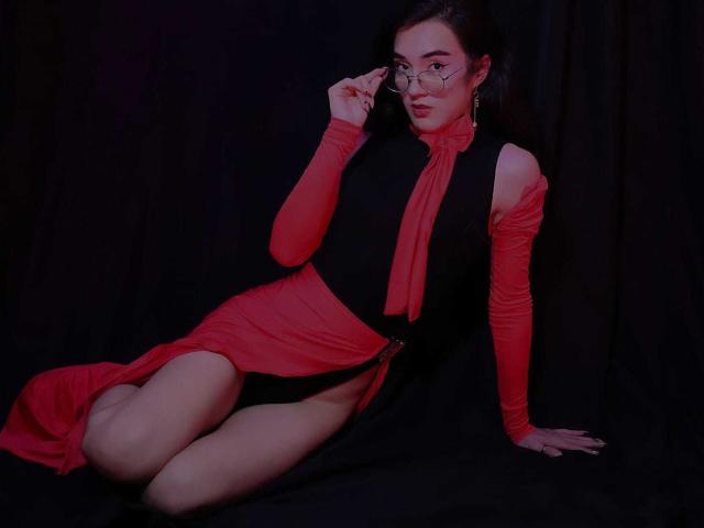 SofiaMonseur in a black dress with red sleeves and skirt, wearing glasses, sitting with one leg up, showing her bare ass and red side slit, against a dark background.