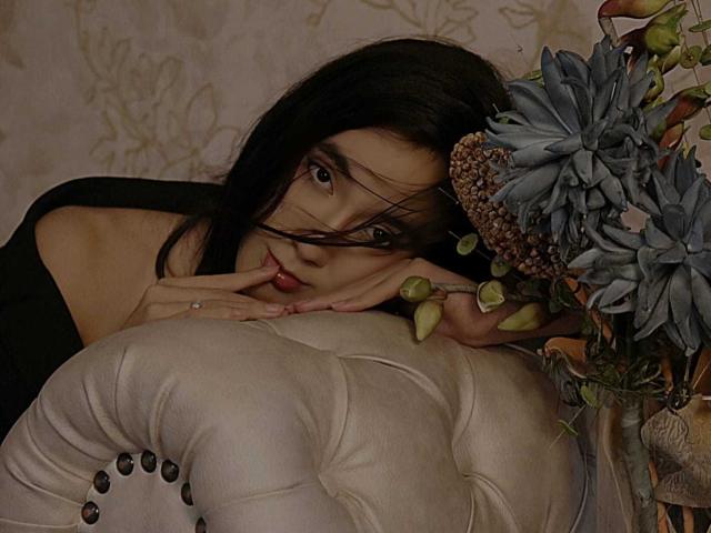 SofiaMonseur with dark hair, black top, fingers near mouth, leaning on white tufted couch, blue succulent flowers, pine cone, and green leaves on right.