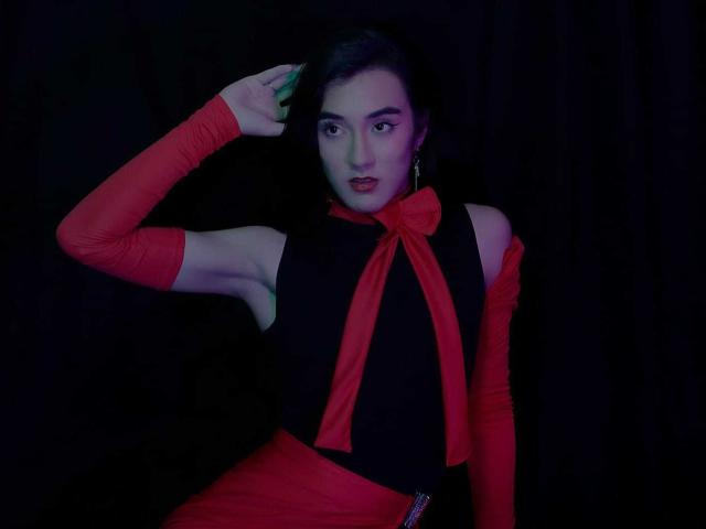 SofiaMonseur with dark hair, red bowtie, red sleeves, black top, and red skirt, touching her head, looking at camera, in dim blue-purple light.