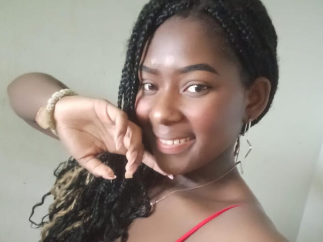 KarinaFlowers, dark-skinned with braided hair, smiling at camera, red strap visible, gold bracelet, silver earrings, hand near face, showing teeth, soft light, cute, confident.