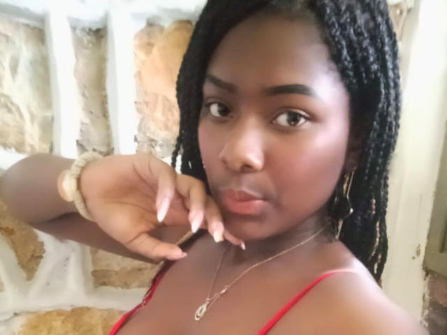 KarinaFlowers with dark skin, black braided hair, red top, gold hoop earrings, and a scrunchie on her wrist, pouting with her finger near her mouth.