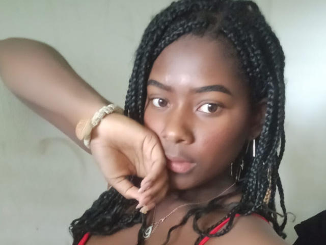 KarinaFlowers with dark skin, black braided hair, and hoop earrings, pouting with her finger near her lips, wearing a red top and a beige bracelet.