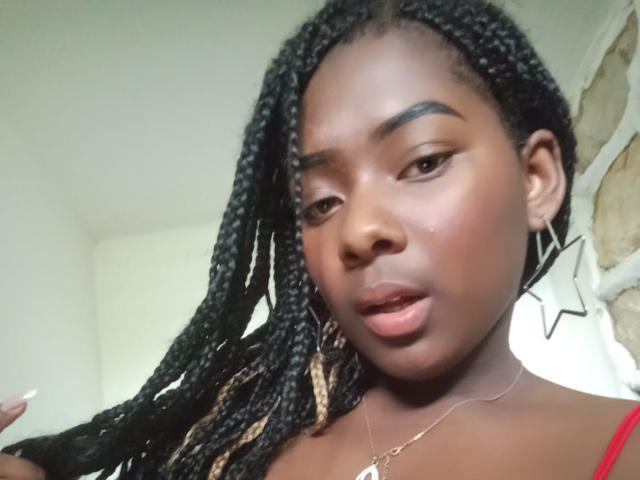 KarinaFlowers has dark skin, long black braided hair with blonde tips, and is wearing a red top, star earrings, and a necklace. Her lips are slightly parted.