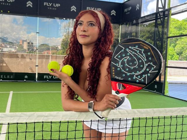 Mia Bianchi, con cabello rojo rizado, sin ropa, sosteniendo una pelota de tenis amarilla y una raqueta de tenis negra y roja, de pie en una cancha de tenis, falda blanca, reloj de metal, red al frente..