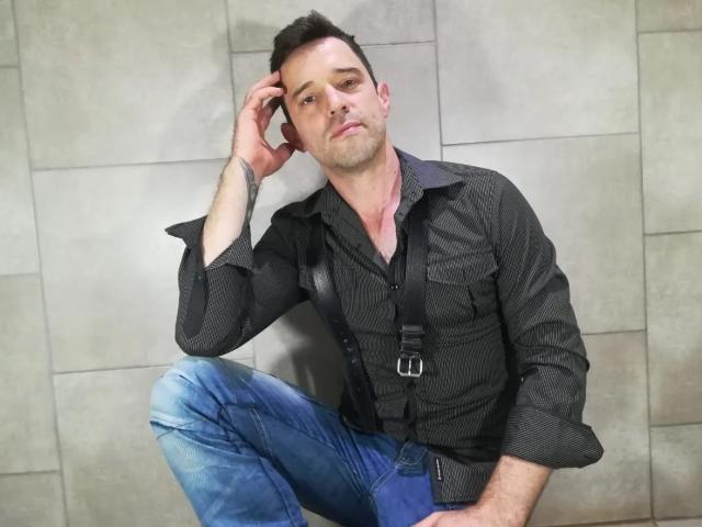 TopBottom is a dude with short brown hair, wearing a black shirt, blue jeans, and black suspenders, sitting with one hand on his head, looking slightly annoyed.