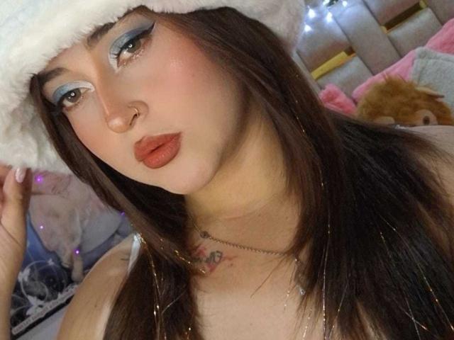 Jheriiz with long brown hair, blue eyeliner, pink blush, red lipstick, white furry hat, nose ring, and necklace, looking at camera, showing off small tits.