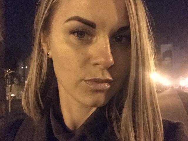 BelleLisaG with shiny, slightly parted lips and a serious expression, wearing a black shirt, has blonde hair, small black earrings, and a slightly visible nose. Nighttime selfie.