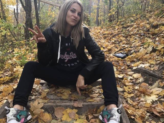 BelleLisaG, sitting on stairs, legs spread, black jacket, black hoodie, black pants, white sneakers, blonde hair, making a peace sign, forest with yellow leaves, autumn.