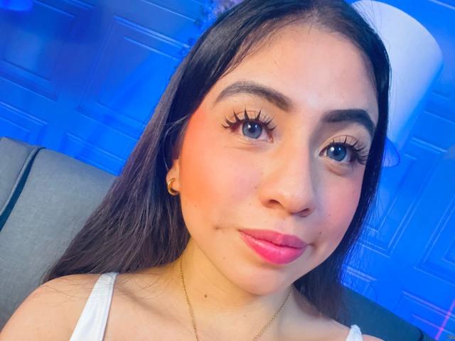ZoeRedds has long brown hair, pink lipstick, and big blue eyes with long lashes. She's wearing a white top and gold necklace, with a subtle smile. Blue light.
