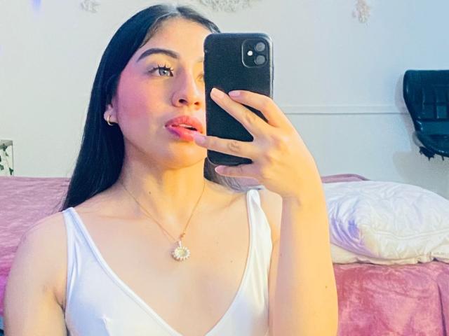 ZoeRedds with long black hair, pouty pink lips, white tank top, holding black phone, necklace with flower pendant, pink bed behind her. Boobs visible, slightly parted.