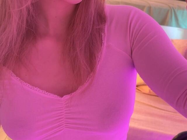 Close-up of Synrix's upper body. She has light pinkish-blonde hair, slightly covering her face. She's wearing a tight, pink, long-sleeve top with a lace-trimmed V-neck, highlighting her medium-sized breasts. The top's fabric is slightly wrinkled, and the lighting casts a pinkish-purple hue over the entire image. The background includes a bed with yellow and green sheets. The composition focuses on the top and her chest area.