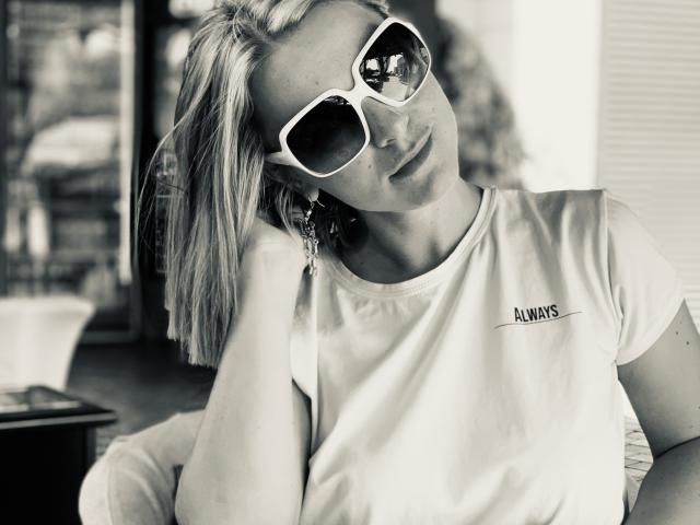 BelleLisaG with blonde hair, wearing white sunglasses and a white tee with "ALWAYS" on it, leaning on her hand, looking slightly to the side, in a black-and-white photo.