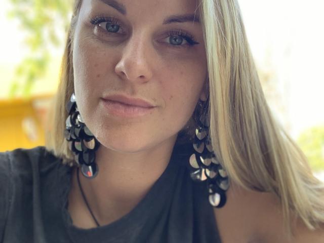 BelleLisaG with blonde hair, blue eyes, and dark earrings featuring black and silver balls. She's wearing a grey top, looking slightly up with a subtle smile.