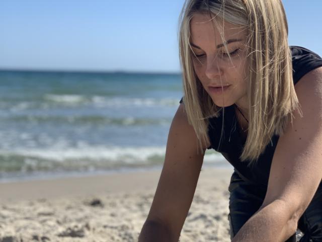 BelleLisaG with blonde hair, black sleeveless top, leaning forward on sandy beach, looking down with a slight smile, ocean and clear sky in background, sunlight on her face.