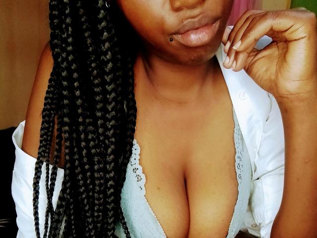 Close-up of EbonyShy-hot's upper torso. She has dark brown skin and braided black hair. She wears a white shirt, open at the chest, revealing a lace-trimmed gray bra that highlights her large breasts. Her left hand is near her chin, with a black lip piercing on her lower lip. The texture of her bra contrasts with the smoothness of her skin and the braids. The image focuses on her chest and facial area. The lighting is natural, highlighting her skin tone and the details of her bra and hair.
